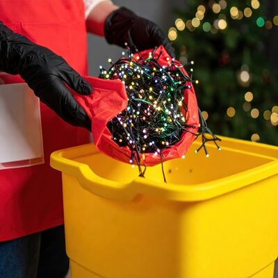 A person wearing black gloves carefully places tangled Christmas lights from a red bag into a yellow container. This depicts the post-holiday cleanup process, focusing on responsible waste disposal and recycling of seasonal decorations after the festive season has ended.